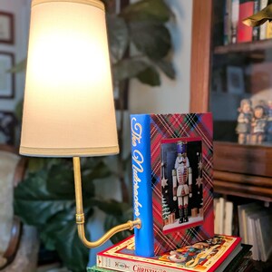 May include: A decorative lamp with a cream lampshade and a brass-toned arm. The lamp is next to a stack of books, including one titled "The Nutcracker" with a festive design featuring a nutcracker figure.