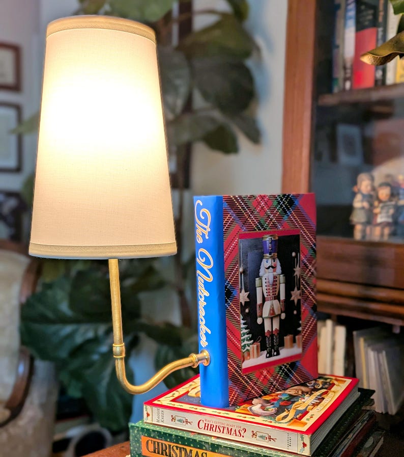 May include: A decorative lamp with a cream lampshade and a brass-toned arm. The lamp is next to a stack of books, including one titled "The Nutcracker" with a festive design featuring a nutcracker figure.