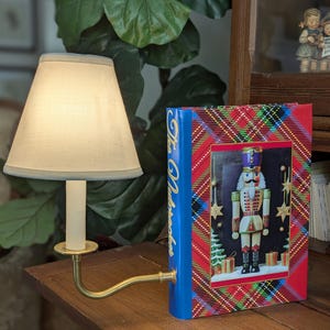 May include: A small brass lamp with a cream lampshade and a decorative book titled "The Nutcracker." The book features a colourful image of a nutcracker soldier on a red and blue plaid background. The lamp is on a wooden surface.