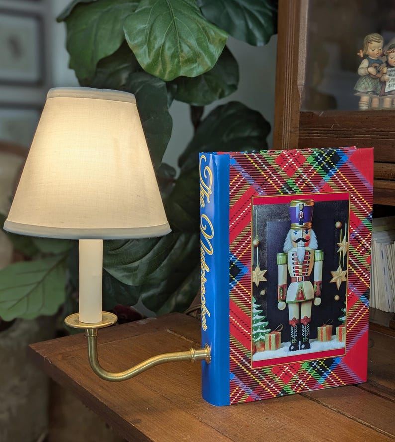 May include: A small brass lamp with a cream lampshade and a decorative book titled "The Nutcracker." The book features a colourful image of a nutcracker soldier on a red and blue plaid background. The lamp is on a wooden surface.