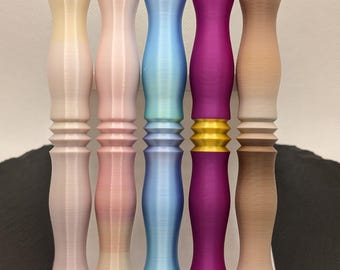 Puede incluir: Una colección de coloridas manijas decorativas en varios tonos de rosa, azul, morado y beige. Las manijas se exhiben en un soporte blanco. El diseño presenta una parte superior redondeada y un cuerpo texturizado y acanalado.