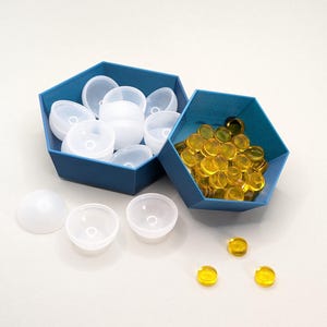 Op de afbeelding: Twee zeshoekige blauwe containers met deksels, een gevuld met witte, koepelvormige plastic objecten en de andere gevuld met gele plastic schijven. Er zijn ook vier witte, koepelvormige plastic objecten en drie gele plastic schijven buiten de containers.