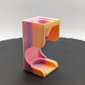 以下が含まれることがあります： ピンク、オレンジ、イエローのグラデーションで3Dプリントされたホルダー。ホルダーの上部に小さな漏斗があり、円形のベースがあります。
