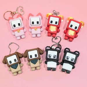May include: A collection of eight colorful keychains featuring blocky animal designs. The keychains include pink cat, red bear, brown dog, and black panda figures. Each keychain has a metal ring for attaching to keys or bags. The background is a solid pink.