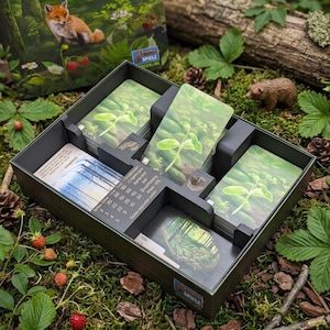 Könnte beinhalten: Eine geöffnete Brettspielbox, die Spielkarten und Komponenten zeigt. Die Box ist dunkelgrün und enthält mehrere Stapel Karten mit Bildern von Pflanzen. Der Hintergrund zeigt eine Waldszene mit einem Fuchs und einem Hirsch.