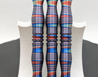 Puede incluir: Tres objetos a rayas en azul, rojo y verde, expuestos sobre un soporte blanco. Los objetos tienen forma cilíndrica con una parte superior e inferior bulbosas. Los objetos tienen un aspecto texturizado y acanalado. El soporte es blanco y curvado.