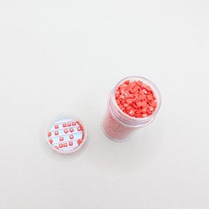 Könnte beinhalten: Ein durchsichtiges Plastikglas gefüllt mit kleinen, quadratischen, korallenfarbenen Perlen, neben einem Deckel mit einem rot-blauen Muster. Die Perlen sind wahrscheinlich für Bastel- oder Kunstprojekte.