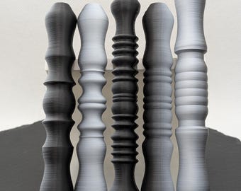 Puede incluir: Seis objetos decorativos, grises y negros, impresos en 3D con diseños variados. Los objetos se exhiben en una estantería blanca sobre un fondo blanco. Los diseños incluyen formas acanaladas, apiladas y bulbosas.