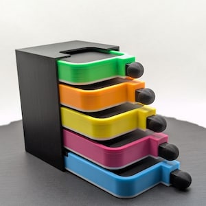 Könnte beinhalten: Ein schwarzer Organizer mit fünf farbigen Schubladen: grün, orange, gelb, pink und blau. Jede Schublade hat einen schwarzen Knopf zum Herausziehen. Die Schubladen sind gestapelt und leicht herausgezogen.
