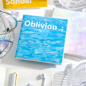May include: A blue notebook with the text "Oblivion" on the cover. The notebook is on a white surface with other notebooks, a pen, and a glass dish.