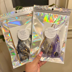 May include: Two clear plastic bags with zipper closures containing moon and star themed spell kits. The bags are iridescent and have a gold sticker on the front with the text "Stay Wild Witchy Craft" and "All In One Full Moon Spell Kit" or "All In One Spiritual Awareness Spell Kit".