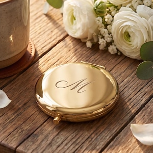May include: A round gold-toned metal compact mirror engraved with the letter M, positioned next to a ceramic mug filled with a warm beverage, a small bouquet of white ranunculus flowers, and a white card with handwritten text.