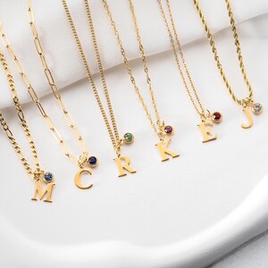 May include: Gold-colored necklaces with various chain styles, each featuring a pendant with a letter and a small gemstone. The letters include M, C, R, K, E, and J. The necklaces are displayed on a white surface.