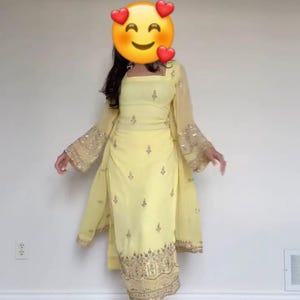 May include: A light yellow salwar kameez suit with gold embroidery. The outfit includes a square-neck top, matching pants, and a sheer shawl with flared sleeves. The sleeves and hem are adorned with gold embellishments. The ensemble is completed with gold-colored sandals.