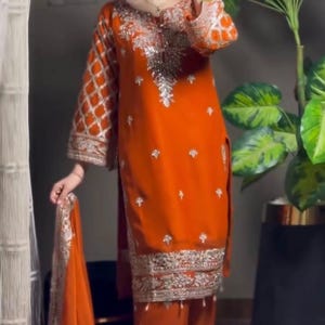 May include: An orange salwar kameez, a traditional South Asian outfit, with silver embroidery on the neckline, sleeves, and hem. The ensemble includes a matching dupatta. The outfit is a vibrant colour.
