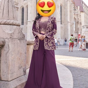 May include: A plum-colored, three-piece outfit featuring a crop top, wide-leg pants, and a long jacket. The jacket and crop top are adorned with gold embroidery. The outfit is worn outdoors in front of a stone building.
