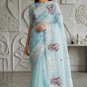 May include: A pale blue saree with delicate white lace edging and floral embroidery. The sheer fabric drapes gracefully, highlighting the detailed design. The saree includes a matching blouse with short sleeves, creating a coordinated and stylish outfit.