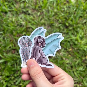 Puede incluir: Dos pegatinas con ilustraciones de perros. Una pegatina representa un perro negro con alas azules, y la otra muestra un perro con un patrón moteado. Las pegatinas se sostienen sobre un fondo de hierba verde.