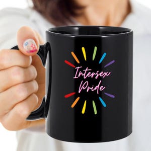 May include: Black ceramic mug with the words "Intersex Pride" in a pink script font, surrounded by colorful rays. The mug has a black handle and is held by a person.