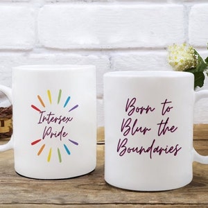 May include: Two white ceramic mugs. One mug has the text "Intersex Pride" with a rainbow burst design. The other mug has the text "Born to Blur the Boundaries" in a cursive font. Both mugs have a curved handle.