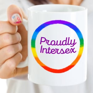 May include: White ceramic mug featuring the text "Proudly Intersex" in purple script, encircled by a rainbow-colored gradient. The mug is held by a person.