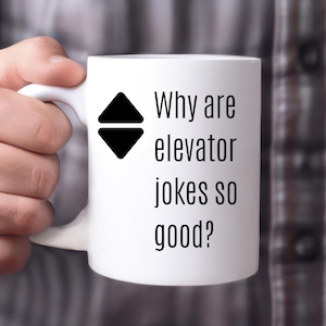 May include: White ceramic mug with a black elevator symbol and the text "Why are elevator jokes so good?" The mug has a curved handle and is held by a person.