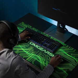 Peut inclure: Un grand tapis de bureau rectangulaire avec un fond noir, un motif de circuit imprimé vert vif et le texte PHANTOMGEARSHOP imprimé en blanc. Le tapis est posé sur un bureau sombre sous un clavier et une souris d'ordinateur noirs.