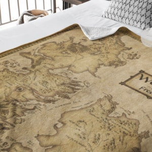 May include: A brown and beige throw blanket with a vintage map of Westeros from the Game of Thrones TV series. The map is printed on the blanket and includes the text "Westeros" and "Free Folk".