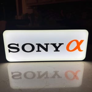 Puede incluir: Un letrero rectangular iluminado que presenta el texto negro SONY y un símbolo alfa naranja. El letrero emite un brillo blanco brillante desde su panel frontal, proyectando un reflejo claro del logotipo sobre la superficie debajo.