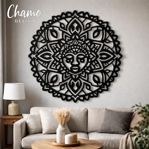 May include: Black, laser-cut wooden mandala wall art with a detailed face design at the center. The mandala is circular with intricate patterns and is mounted on a neutral-colored wall. The text "Chamo Design" is in the upper left corner.