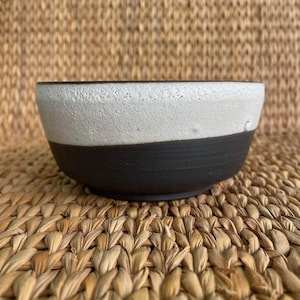 May include: A handcrafted ceramic bowl with a two-tone design. The top half is a textured off-white, while the bottom half is a matte black. The bowl has a rounded shape and sits on a woven surface.