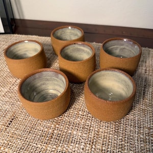 May include: A set of five small ceramic cups with a textured, matte brown exterior and a smooth, light cream-colored interior glaze. Each cup features a simple, handleless cylindrical shape with a slightly rounded base and a wide, open rim.