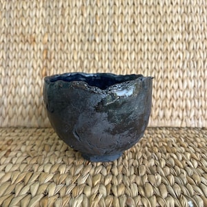 May include: A handcrafted, dark-colored ceramic bowl with a textured surface and a wavy rim. The bowl has a deep interior and a slightly rough exterior, showcasing the unique characteristics of the pottery.