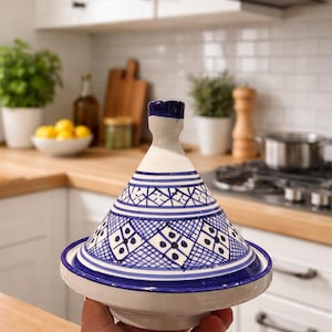 May include: A white and blue ceramic tagine pot with a conical lid. The lid and base feature intricate blue geometric patterns. The tagine is held in front of a kitchen backdrop with white cabinets and wooden countertops.