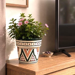 May include: A potted plant with green leaves and small pink flowers in a white ceramic planter. The planter features a hand-painted geometric pattern in dark green, black, and terracotta red, including horizontal lines, dots, and triangular shapes.