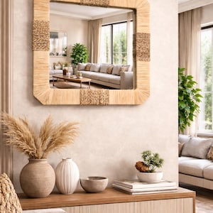 Può includere: Uno specchio quadrato con una cornice in fibra naturale intrecciata è appeso a una parete beige. Lo specchio riflette un soggiorno con un divano, un tavolino e grandi finestre. Sotto lo specchio c'è una consolle in legno con vasi decorativi e una piccola pianta.