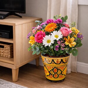 May include: A vibrant floral arrangement in a decorative yellow and black pot. The bouquet features pink roses, white daisies, orange gerberas, and other colorful blooms. The pot has an intricate pattern.