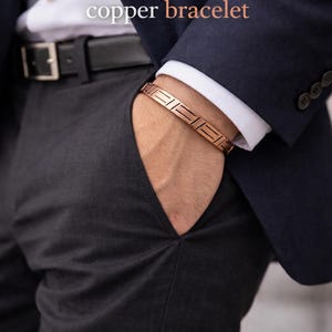 Pode incluir: Elegante pulseira de cobre. Apresenta-se uma pulseira de cobre com um design geométrico. A pulseira é usada num pulso, com um blazer azul escuro e calças cinzentas no fundo. O texto "Elegant copper bracelet" é visível.