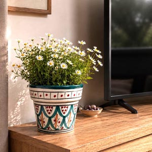 May include: A ceramic flower pot featuring a hand-painted pattern of teal scalloped arches, red dots, and thin orange vertical lines. The pot contains a lush, bushy plant with numerous small white daisy-like flowers with yellow centers.