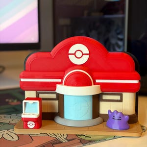 May include: A 3D-printed model of a Pokémon Center, with a red roof, white Poké Ball symbol, and beige walls. It includes a blue door, a small red vending machine, and a purple Ditto figure. The model is set on a tan base.