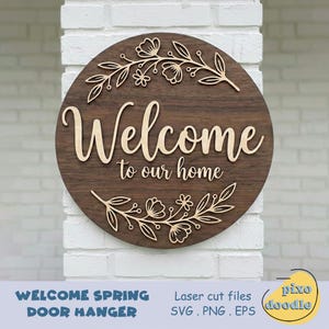 Puede incluir: Un colgador de puerta redondo de madera con las palabras "Welcome to our home" en madera clara sobre un fondo de madera oscura. Diseños florales cortados con láser en la parte superior e inferior. Las palabras "Welcome Spring Door Hanger" están en la parte inferior.