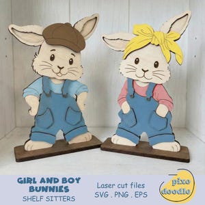 May include: Two wooden bunny shelf sitters. One bunny wears a brown cap, blue overalls, and a light blue shirt. The other bunny wears a yellow headband, blue overalls, and a pink shirt. Both are on brown bases. Text reads "Girl and Boy Bunnies Shelf Sitters".
