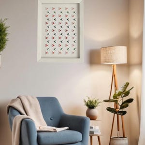 May include: A framed artwork with a white border hangs on a wall. The artwork features a repeating pattern of red, green, and black triangles on a white background. The room includes a blue armchair, a floor lamp, and a potted plant.