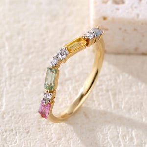 Op de afbeelding: Een gouden ring met een rij kleurrijke edelstenen. De ring heeft een delicaat ontwerp met rechthoekig geslepen stenen in roze, groen, geel en blauw, afgewisseld met kleine, ronde, heldere stenen. De band is van gepolijst goud.