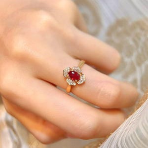 May include: A gold ring with a floral design. It has a central oval-cut, deep red gemstone, encircled by a halo of small, clear stones. The ring is set on a slender gold band.