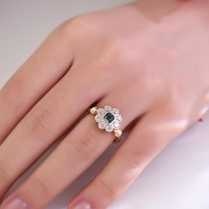 May include: A gold ring featuring a central square-cut, teal-colored gemstone surrounded by a halo of smaller, clear gemstones. The ring's design resembles a flower, with the gemstones set in a floral pattern. The band is a simple, polished gold.