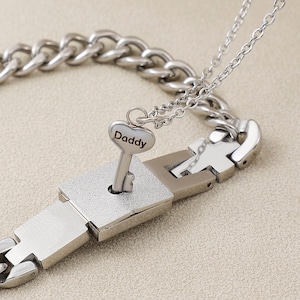 Pode incluir: Um conjunto de colar e pulseira de prata com um design de cadeado e chave. A chave tem um pingente em forma de coração com a palavra "Pai" gravada.