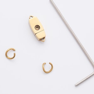 Peut inclure: Ensemble de composants pour la fabrication de bijoux, comprenant un fermoir doré, deux petits anneaux dorés en forme de C et une clé Allen argentée. Les articles sont disposés sur un fond blanc, suggérant un thème artisanal ou de réparation.