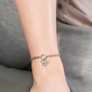 May include: A silver-colored anklet with a delicate bow charm. The anklet is made of small, linked chain segments and is worn on the ankle. The bow charm is centered and adds a touch of elegance to the jewelry.