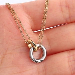 Puede incluir: Un collar de cadena dorada con un colgante de doble anillo. Los anillos están entrelazados y tienen las palabras "Daddy" y "Good Girl" grabadas.